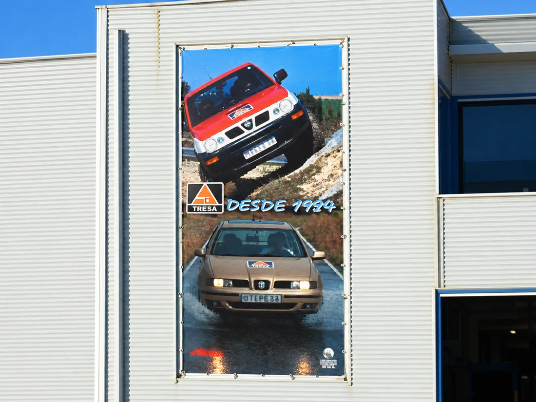 Lona de gran formato de TRESA instalada en la fachada metálica de una nave, con imágenes de vehículos y el lema DESDE 1994.